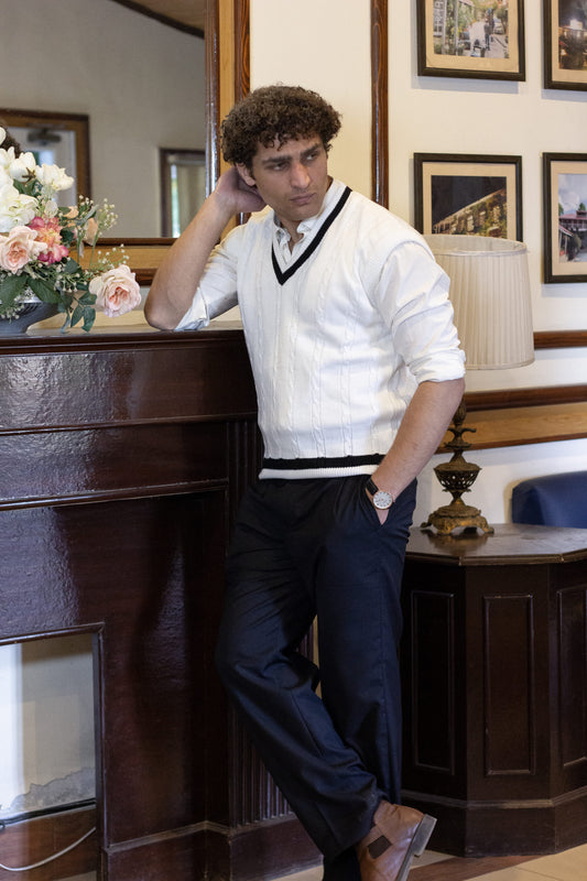 Man wearing a White Cable knitted Sleeveless sweater over a white shirt and black pants in an vintage old money setting showing full outfit